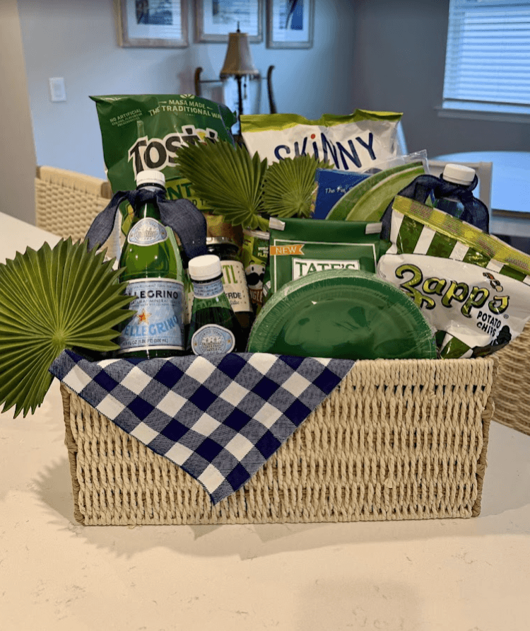 Green-themed welcome basket with Tostitos, Pellegrino, Tate's cookies, Zapp's chips, a green plate, and a navy gingham napkin